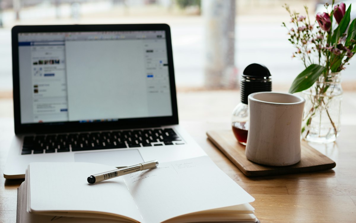 A laptop, notebook and coffee on a bright desk.