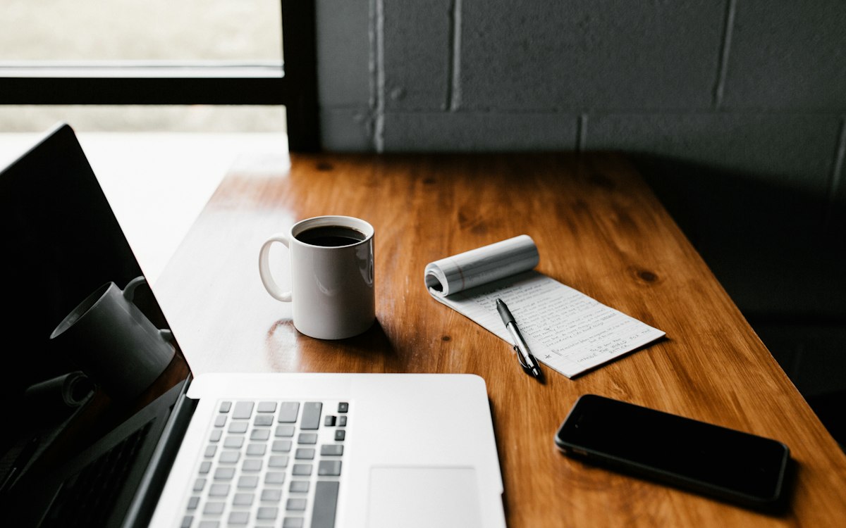 A laptop, coffee, phone and notepad on a wooden desk.